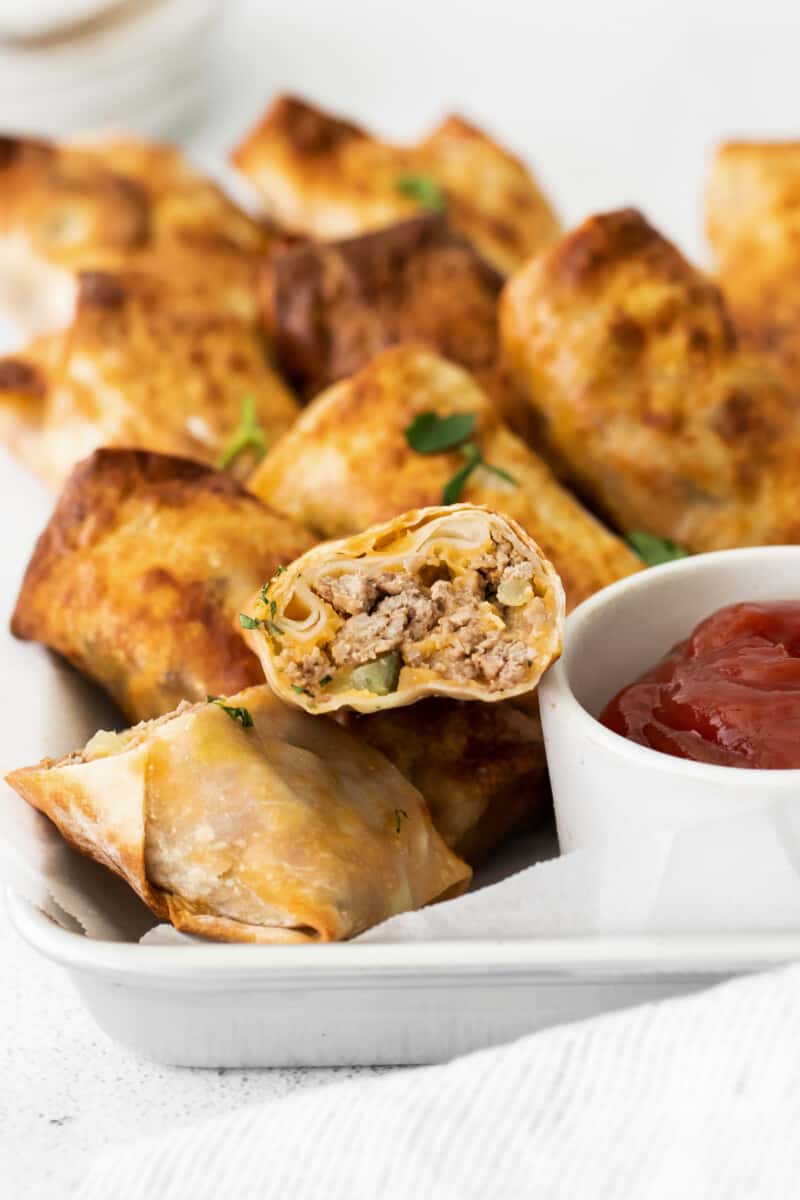 cheeseburger egg rolls with one cut in half showing the filling on a serving tray with a side of ketchup in a white bowl
