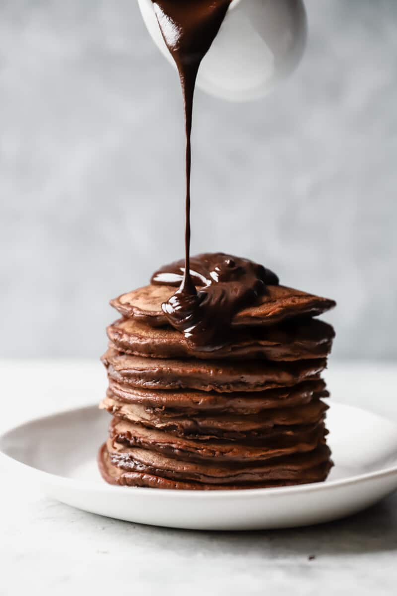 pouring chocolate sauce onto a stack of chocolate pancakes on a white plate
