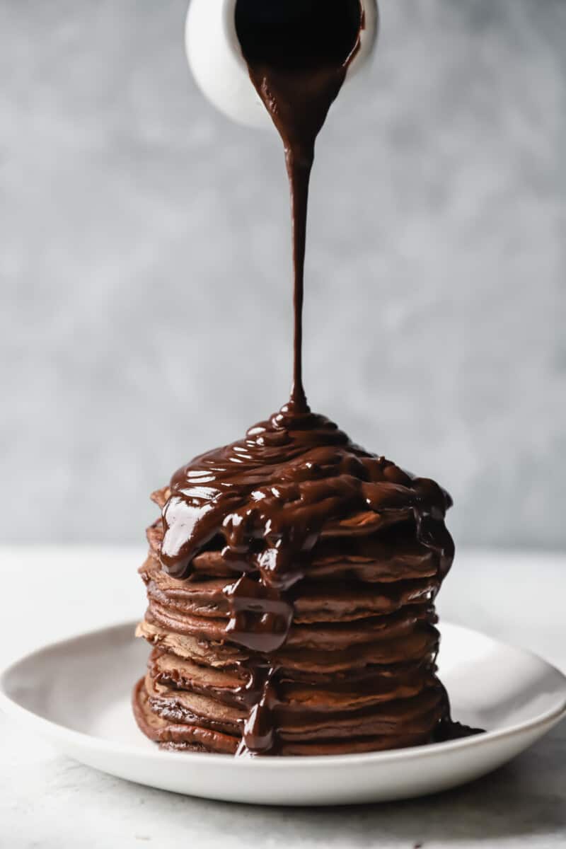 pouring chocolate sauce onto a stack of chocolate pancakes on a white plate