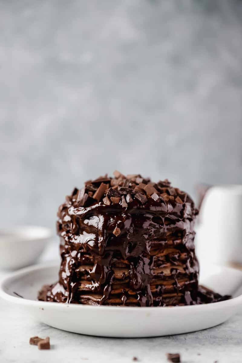 stack of chocolate pancakes topped with chocolate sauce and chopped chocolate on a white plate