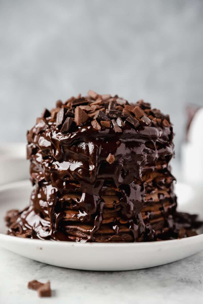 stack of chocolate pancakes topped with chocolate sauce and chopped chocolate on a white plate