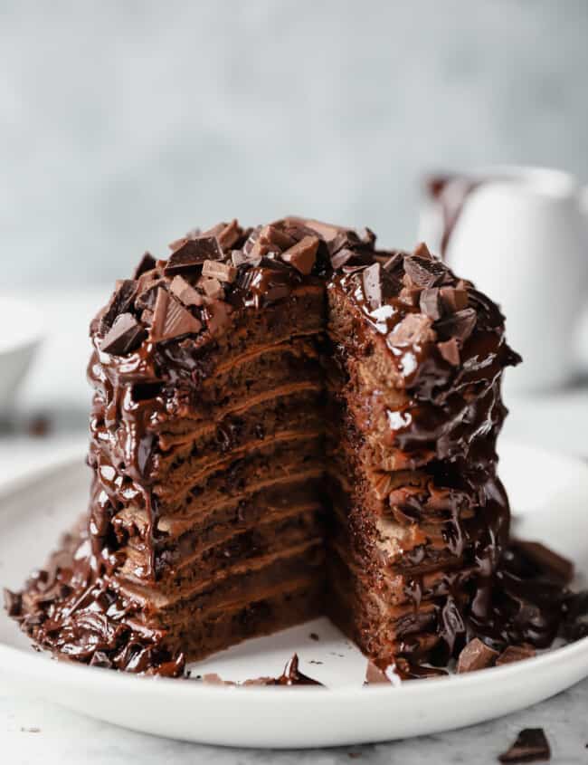 stack of chocolate pancakes topped with chocolate sauce and chopped chocolate on a white plate
