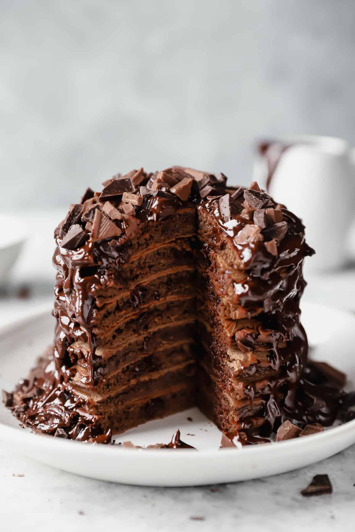 stack of chocolate pancakes topped with chocolate sauce and chopped chocolate on a white plate