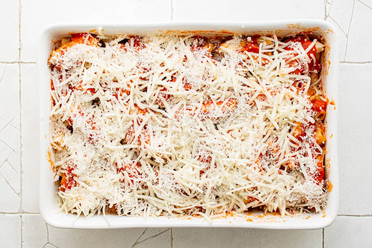 shredded cheese on top of noodles, sauce, and chicken in a white baking dish