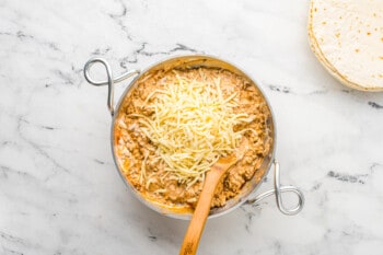 mixing shredded cheese into beef filling in a saucepan with a wood spoon
