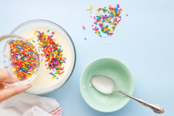 hand pouring bowl of sprinkles into melted white chocolate and coconut oil mixture in a glass bowl