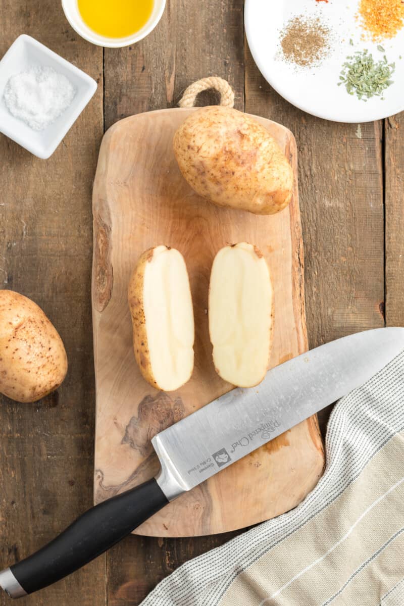 Potato sliced in half on a wooden cutting board with a knife, olive oil and spices in the background.