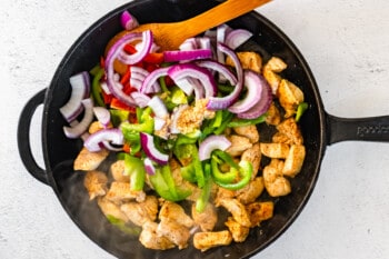 sliced onion and bell pepper added to cooked chicken in a skillet with a wood spoon