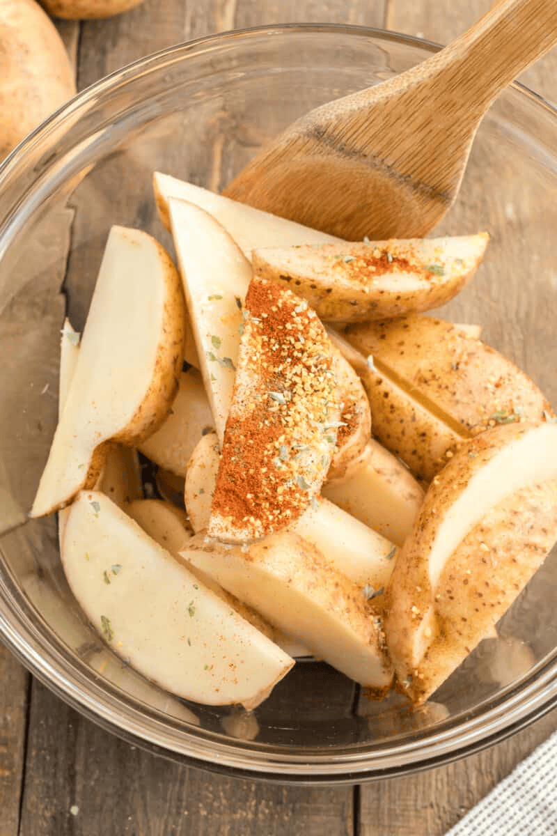 Seasoned potato wedges in a glass bowl with a wooden spoon.