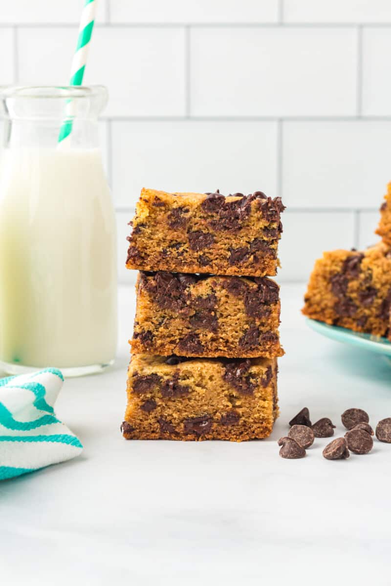 3 chocolate chip cookie bars stacked next to a glass of milk.