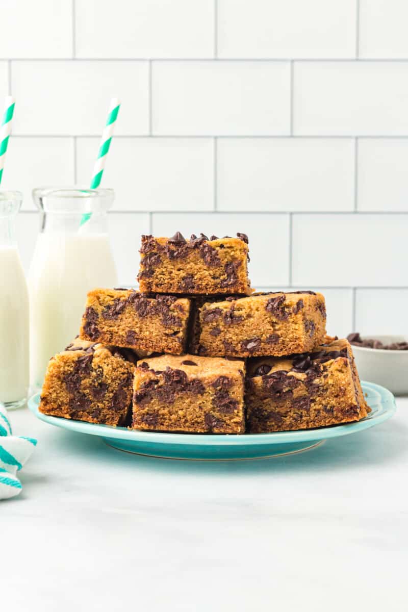 chocolate chip cookie bars stacked on a blue plate with glasses of milk.