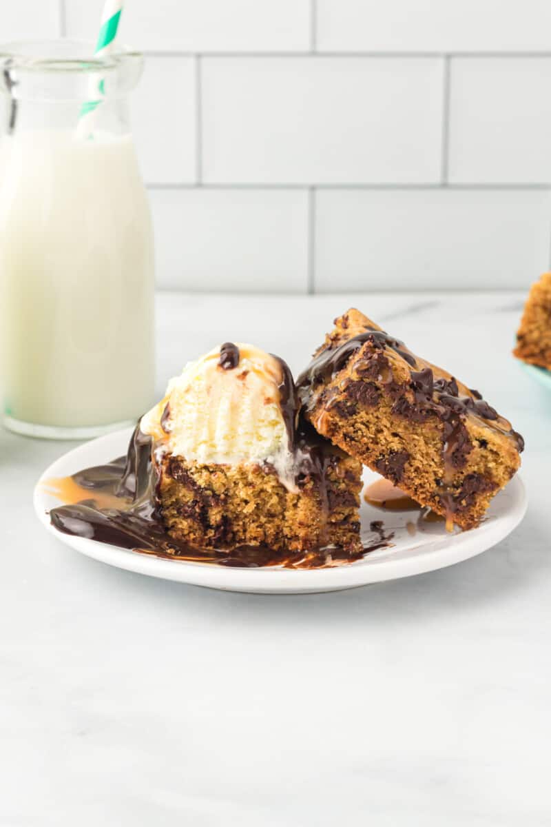 2 chocolate chip cookie bars topped with ice cream and chocolate sauce on a white plate.