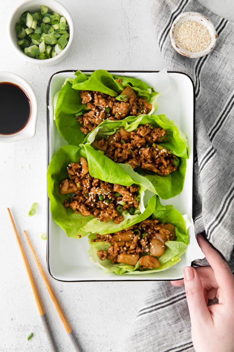 a hand grabbing a white tray of pf changs lettuce wraps with chopsticks.