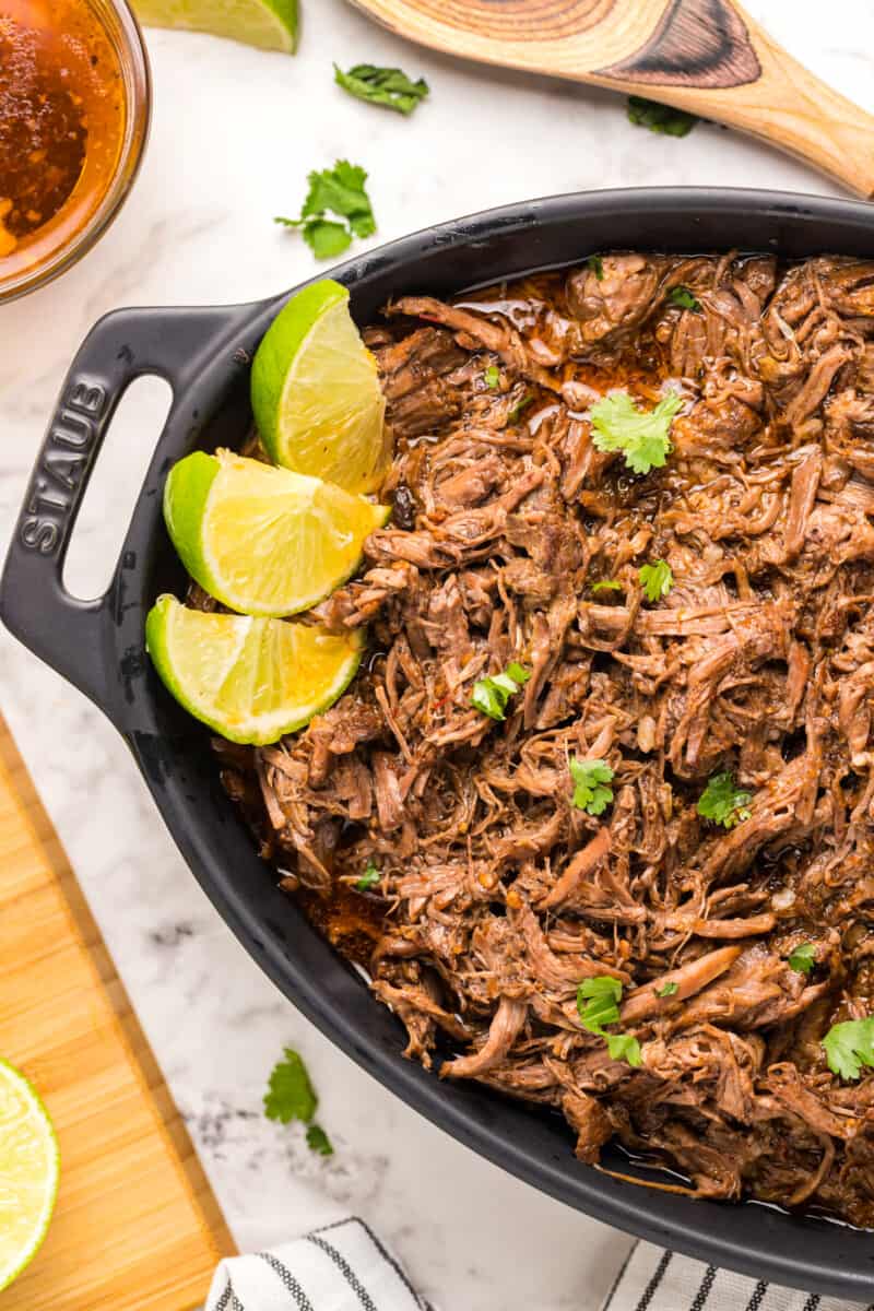 close up of barbacoa beef in a cast iron serving vessel with limes.
