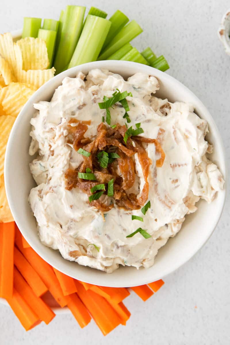 overhead image of french onion dip in a white bowl on a serving platter surrounded by chips, celery sticks, and carrot sticks