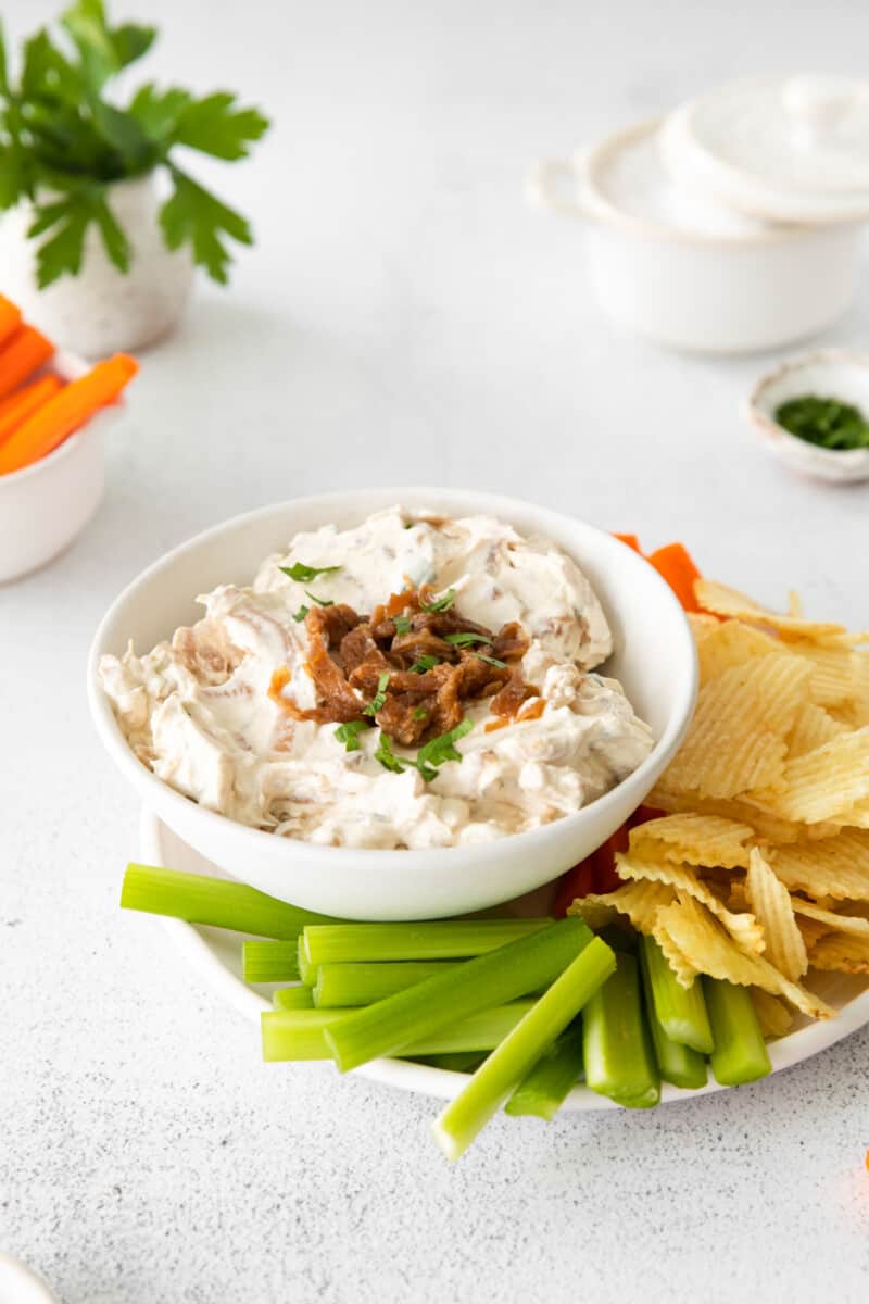 french onion dip in a white bowl on a serving platter surrounded by chips, celery sticks, and carrot sticks