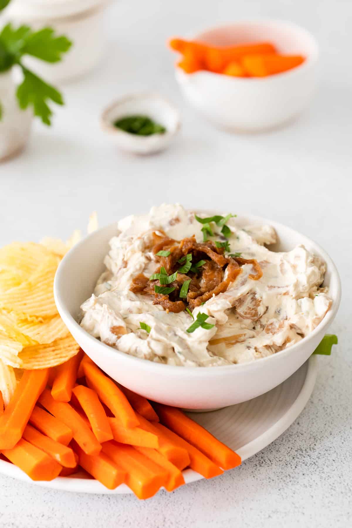 french onion dip in a white bowl on a serving platter surrounded by chips, celery sticks, and carrot sticks