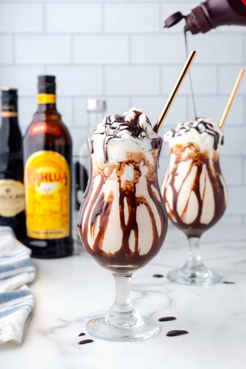 2 frozen mudslides with gold straws drizzled with chocolate syrup in front of bottles of alcohol.