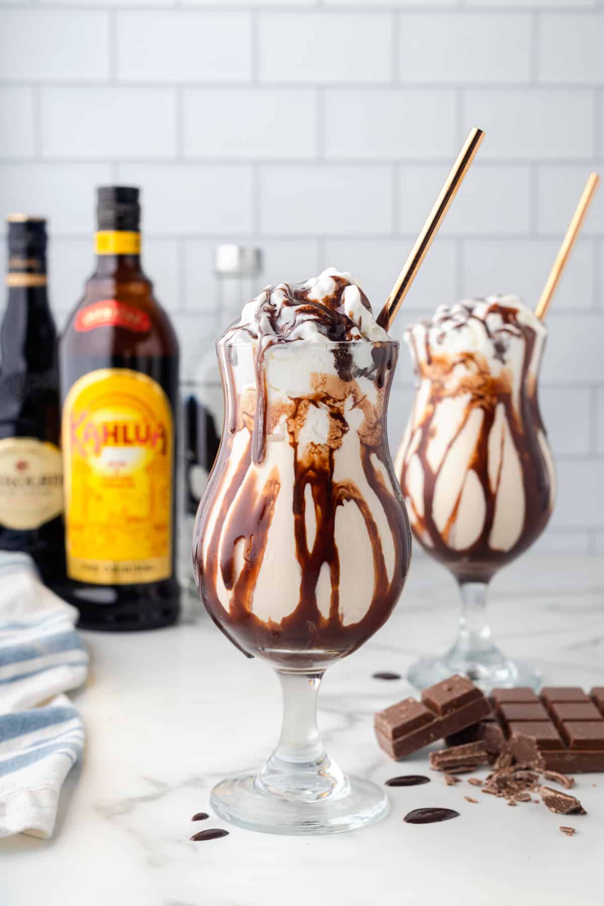 2 frozen mudslides with gold straws in front of bottles of alcohol.