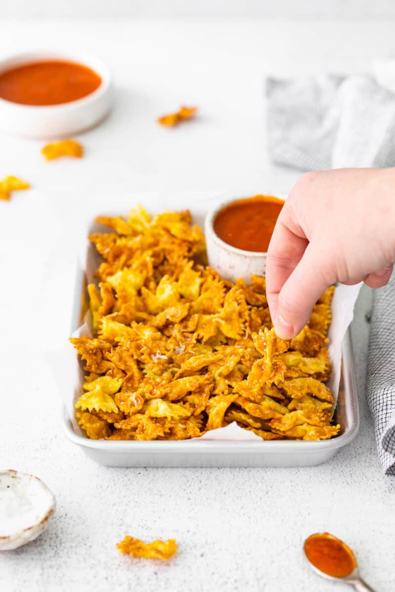 a hand grabbing a garlic parmesan pasta chip from a tray with marinara sauce.