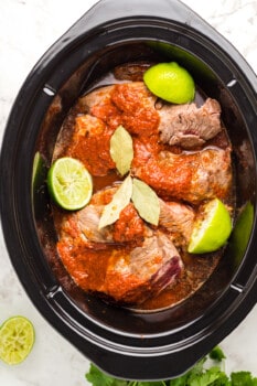 chuck roast in a crockpot with marinade, limes, and bay leaves.