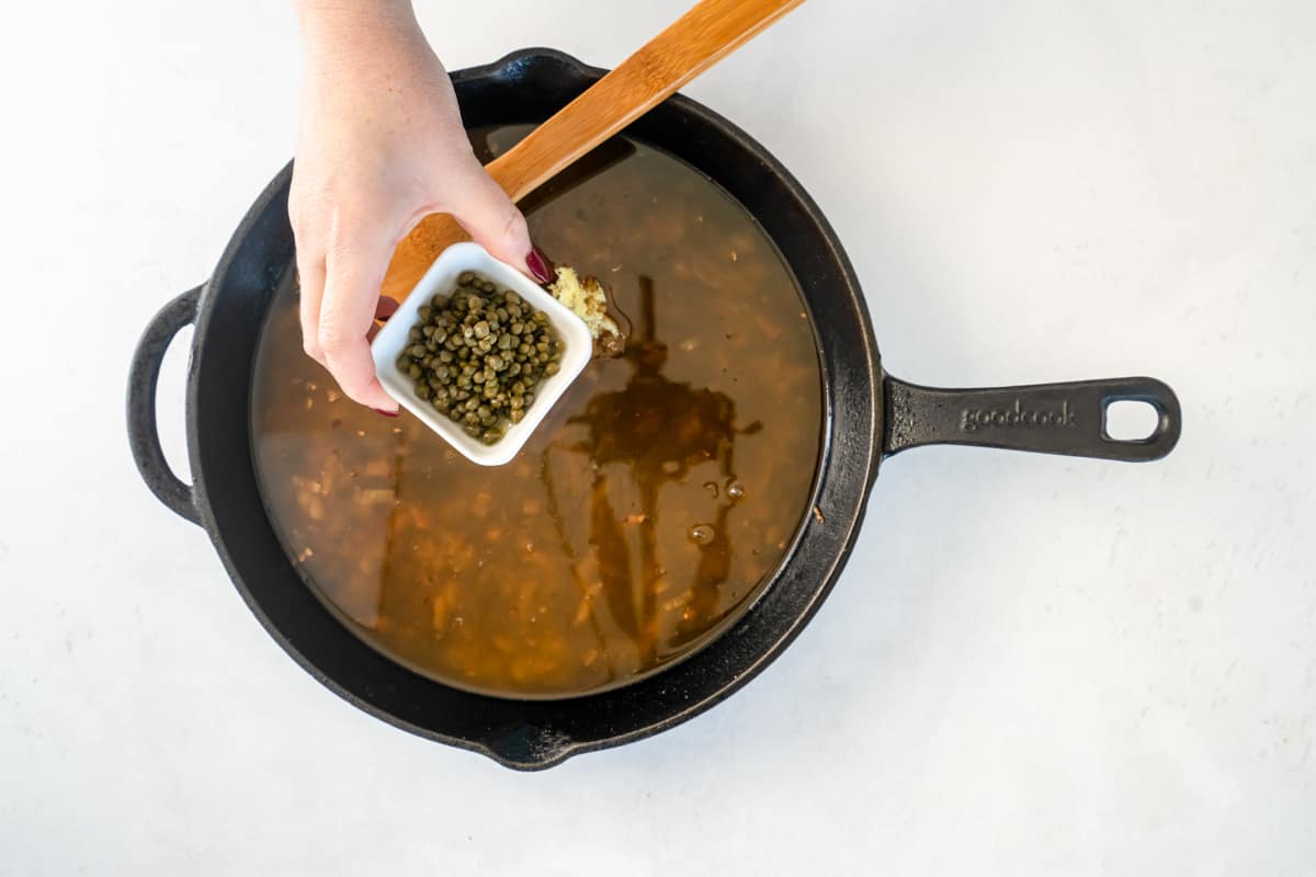 Overhead view of capers being added to a skillet with a wooden spoon.