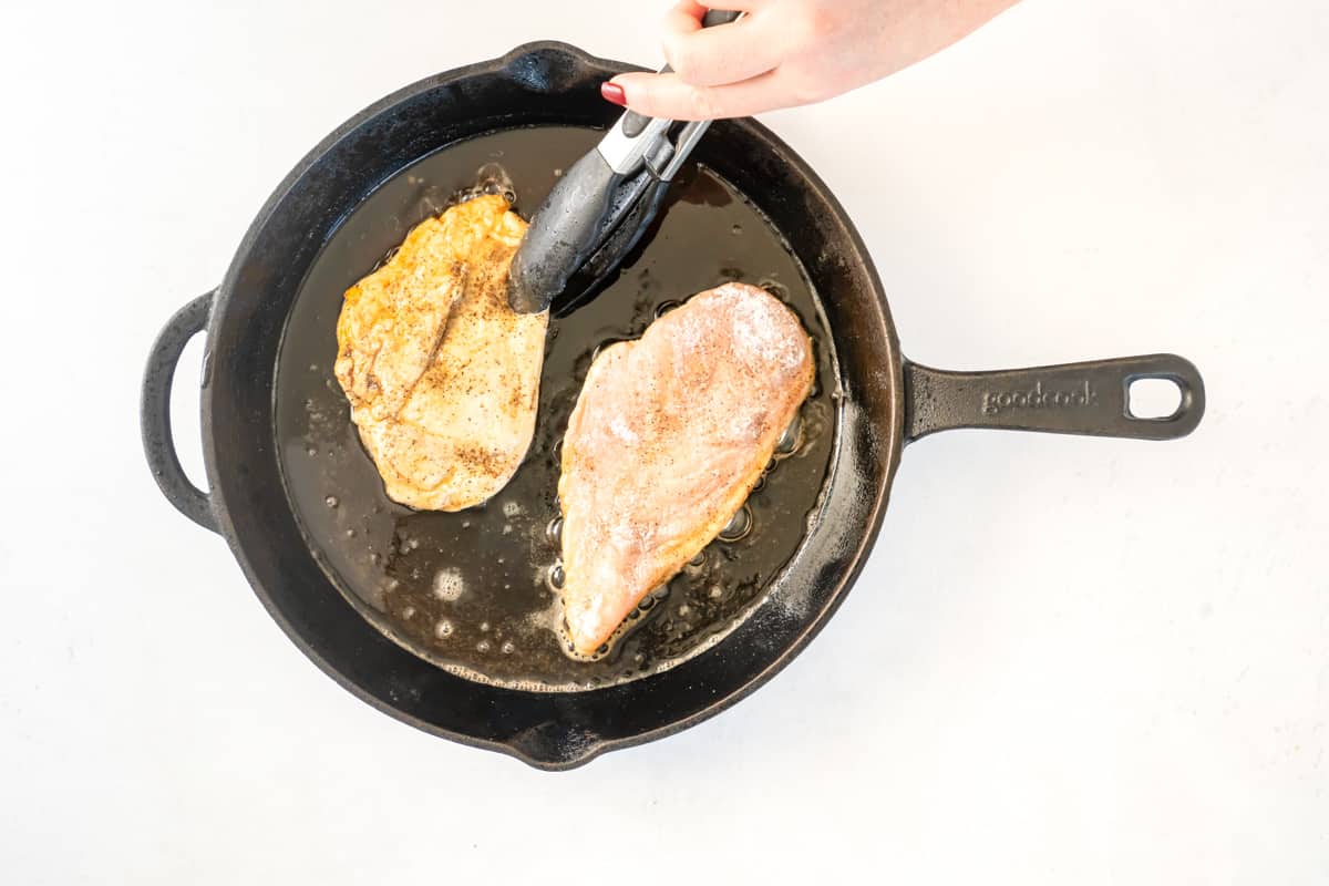 Overhead view of chicken being cooked in a skillet with tongs.