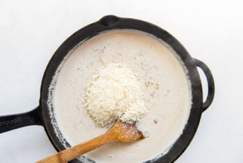 Parmesan cheese added to cream sauce in a skillet with a wooden spoon.