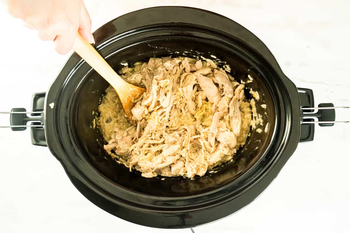 Shredded chicken being stirred with a wooden spoon into the crockpot.