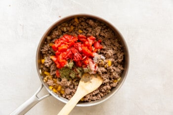 seasonings and tomatoes added to ground beef mixture in a skillet with a wood spoon