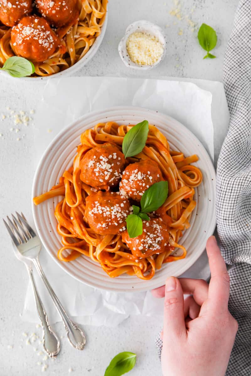 overhead view of a hand grabbing chicken parmesan meatballs over spaghetti in a white bowl with 2 forks.
