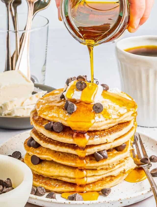 syrup poured over a tall stack of gluten free pancakes on a white plate with chocolate chips and whipped cream.