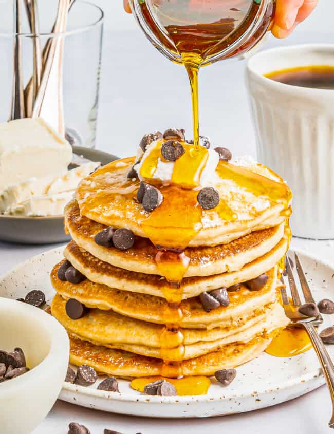 syrup poured over a tall stack of gluten free pancakes on a white plate with chocolate chips and whipped cream.