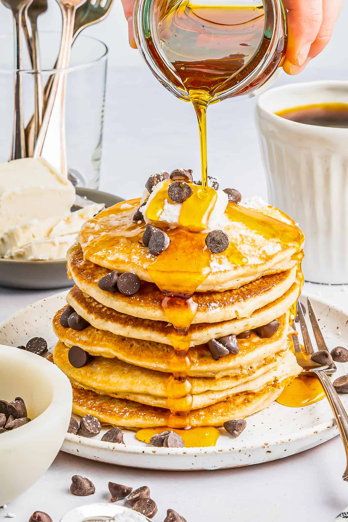 syrup poured over a tall stack of gluten free pancakes on a white plate with chocolate chips and whipped cream.