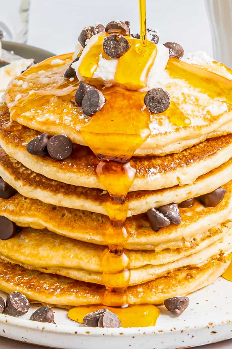 a tall stack of gluten free pancakes on a white plate with chocolate chips maple syrup and butter.