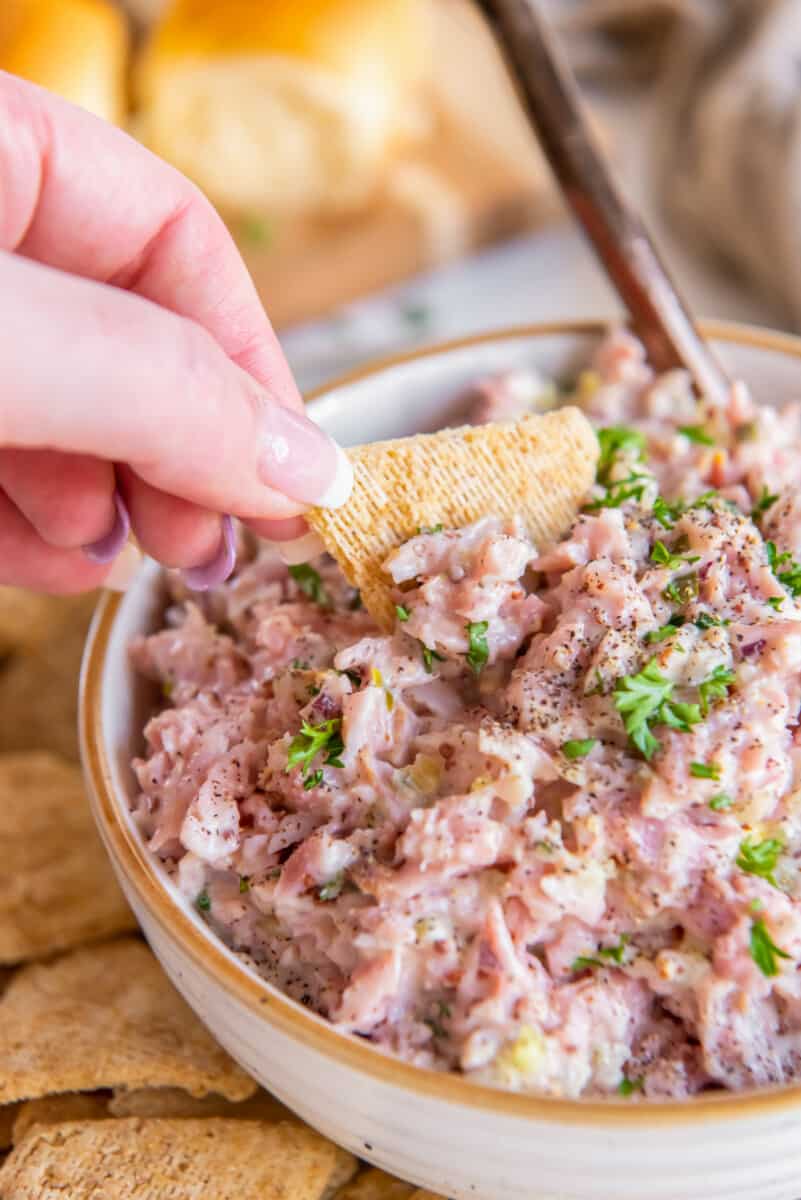 a hand dipping a triscuit into a bowl of ham salad.