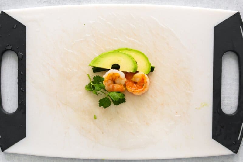 shrimp avocado cucumber and cilantro on rice paper on a cutting board.