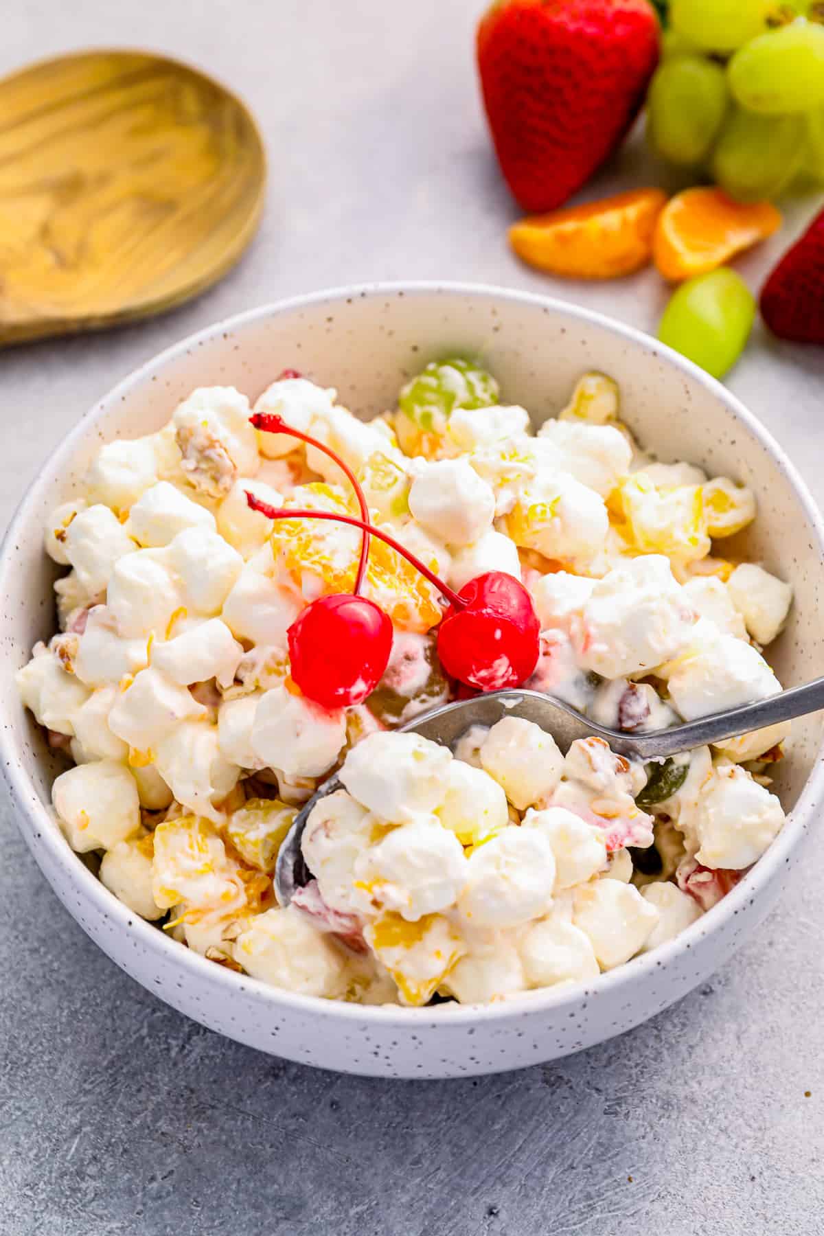 ambrosia salad in a white bowl with 2 maraschino cherries and a spoon.