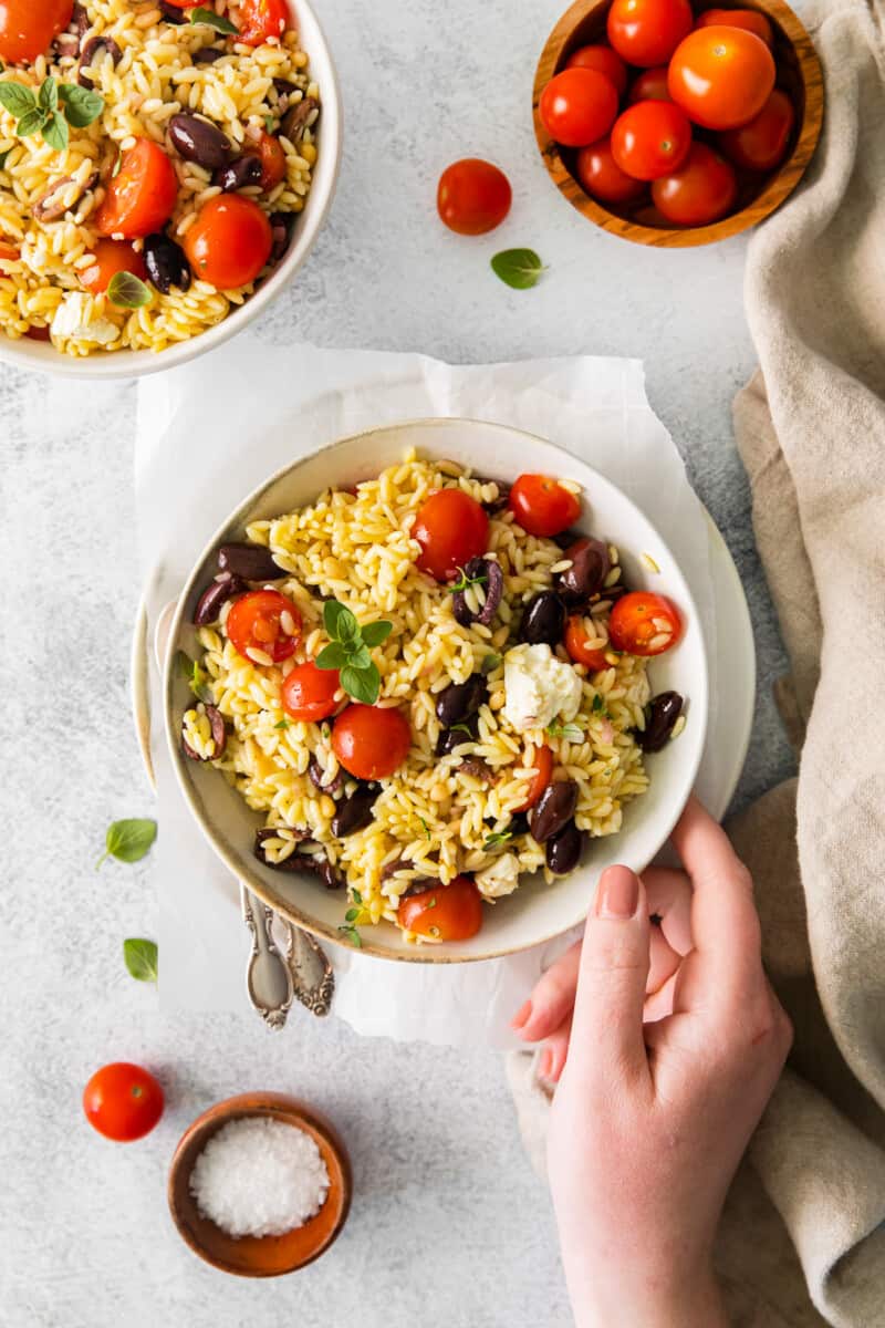 a hand grabbing mediterranean orzo salad in a white bowl.