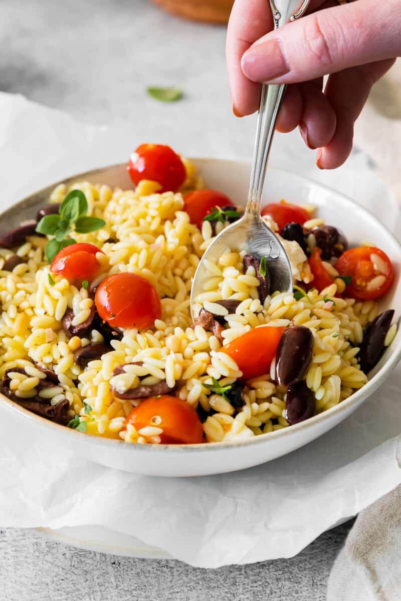 a spoonful of mediterranean orzo salad hovering over mediterranean orzo salad in a white bowl.