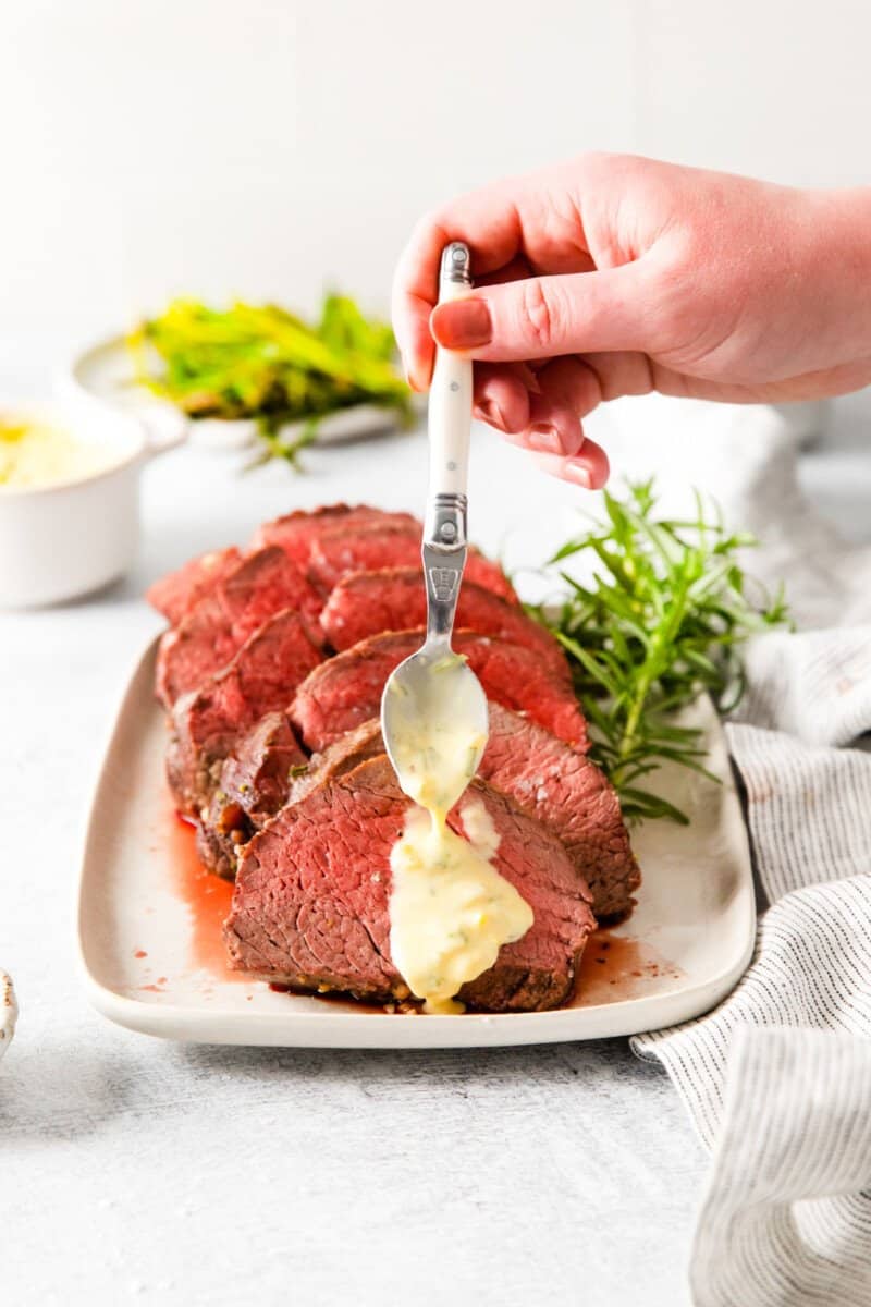 spoon pouring Bearnaise sauce over crockpot beef tenderloin on a white plate.