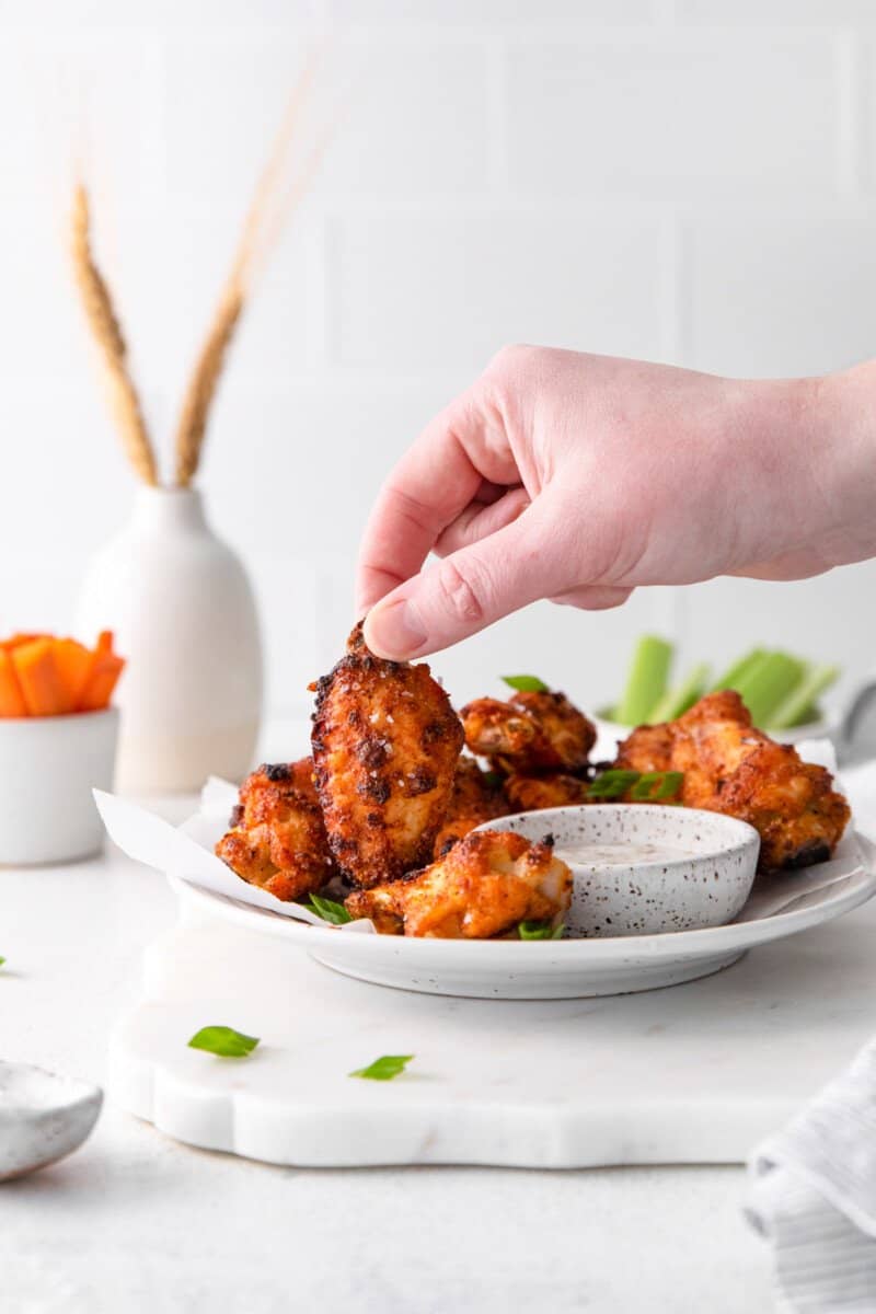 hand grabbing a dry rub chicken wing from a pile of wings on a white plate with dipping sauce.