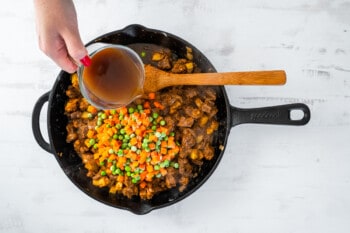 beef broth carrots and peas added to beef mixture in a skillet with a wooden spoon.