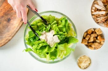 chicken caesar salad dressing on romaine lettuce in a glass bowl with tongs.