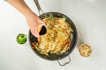 soy sauce added to chicken lo mein in a frying pan.
