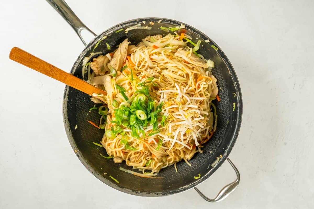 Adding bean sprouts and green onions to the chicken lo mein in a frying pan with a wooden spoon. - 13