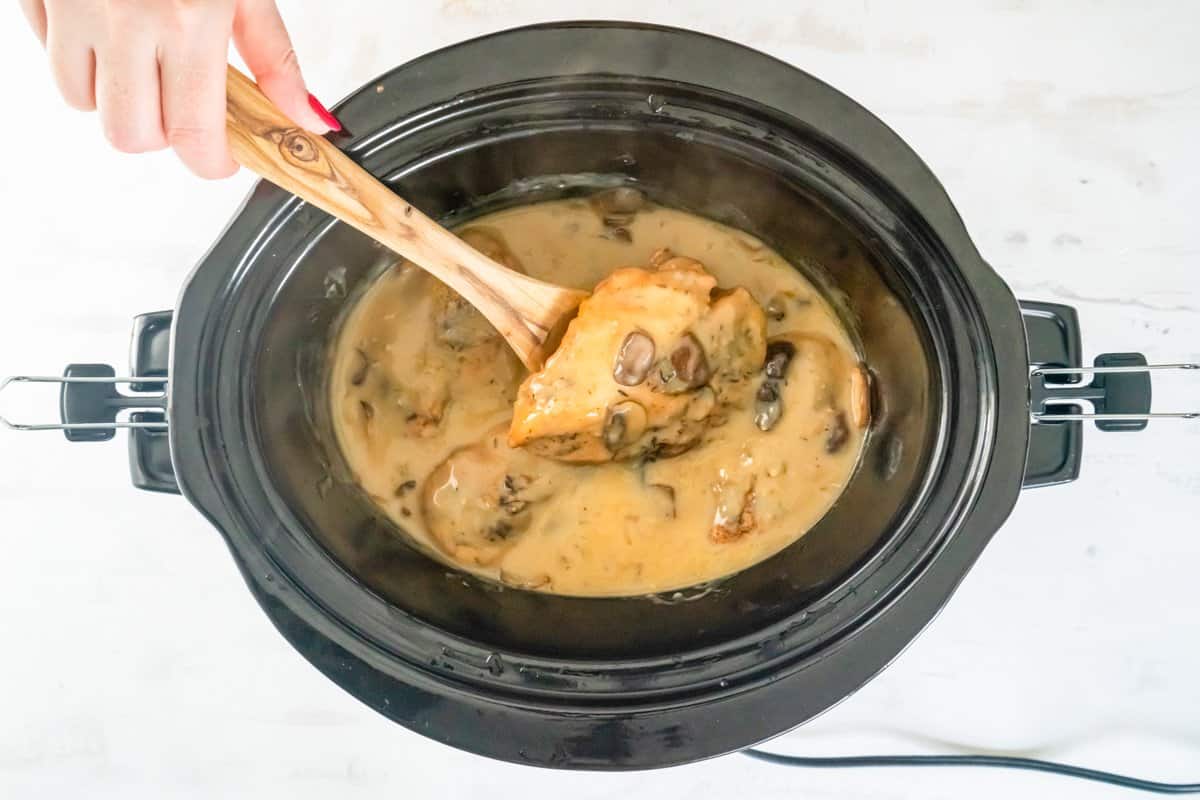 a wooden spoon lifting a chicken breast out of crockpot chicken marsala.