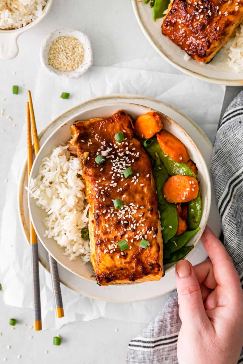 hand grabbing air fryer miso salmon in a white bowl with rice carrots and snap peas.