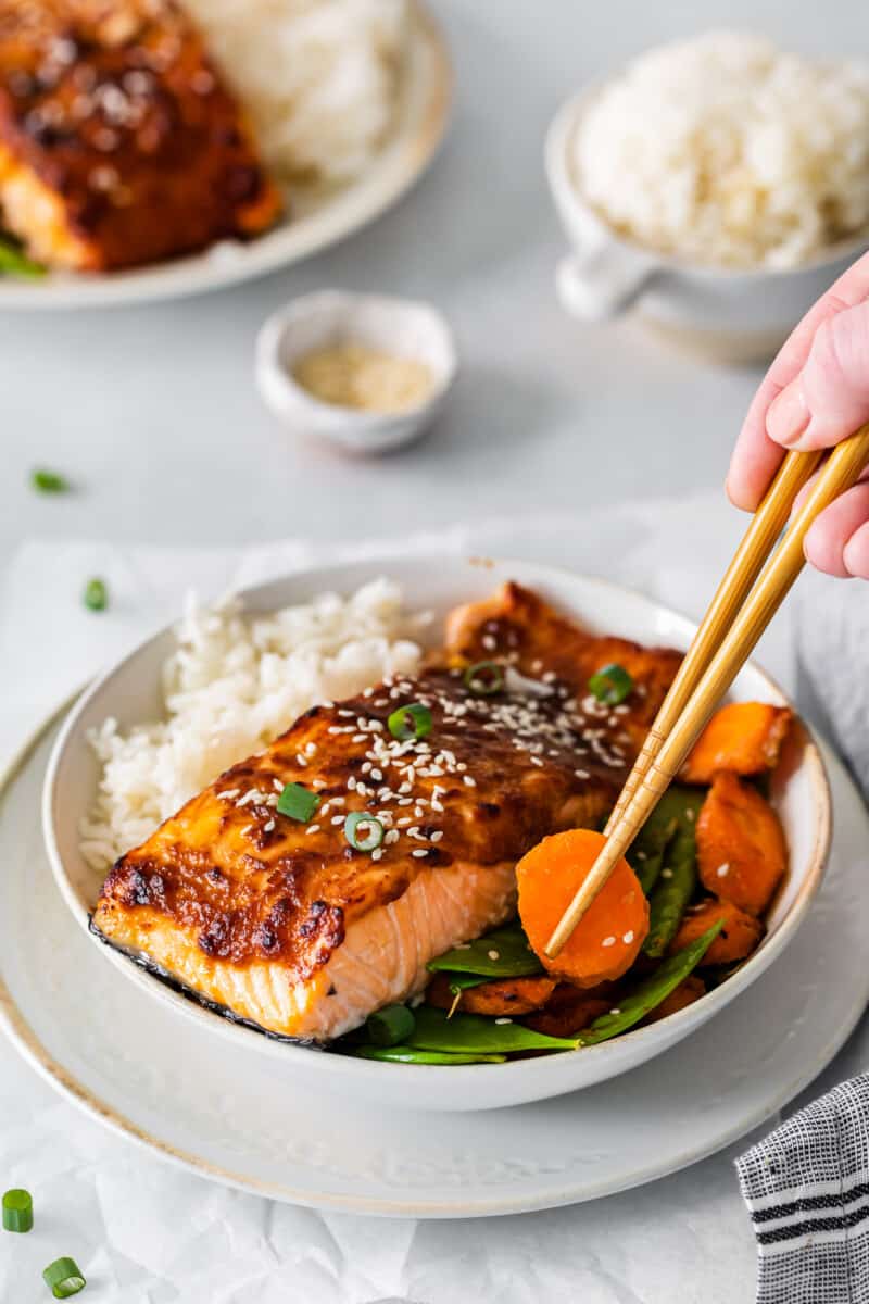 chopsticks grabbing a carrot from a bowl of air fryer miso salmon with rice carrots and snap peas.
