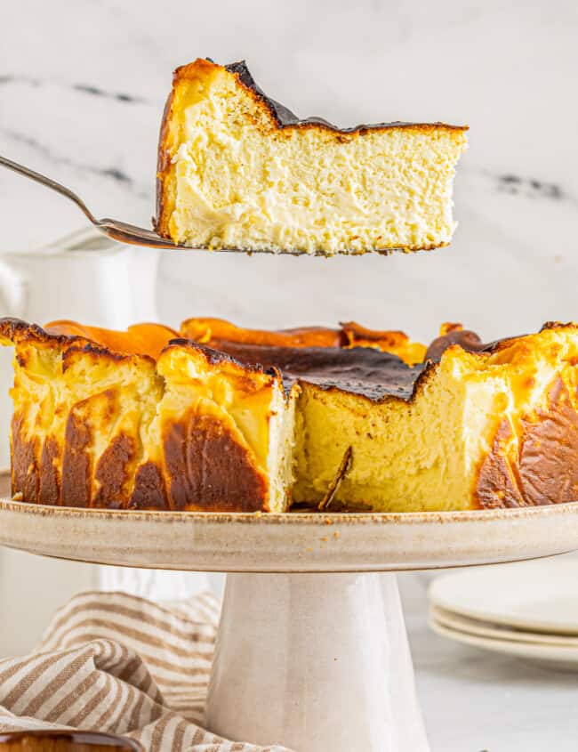 a slice of basque cheesecake on a cake server hovering above a cheesecake on a white cake stand.